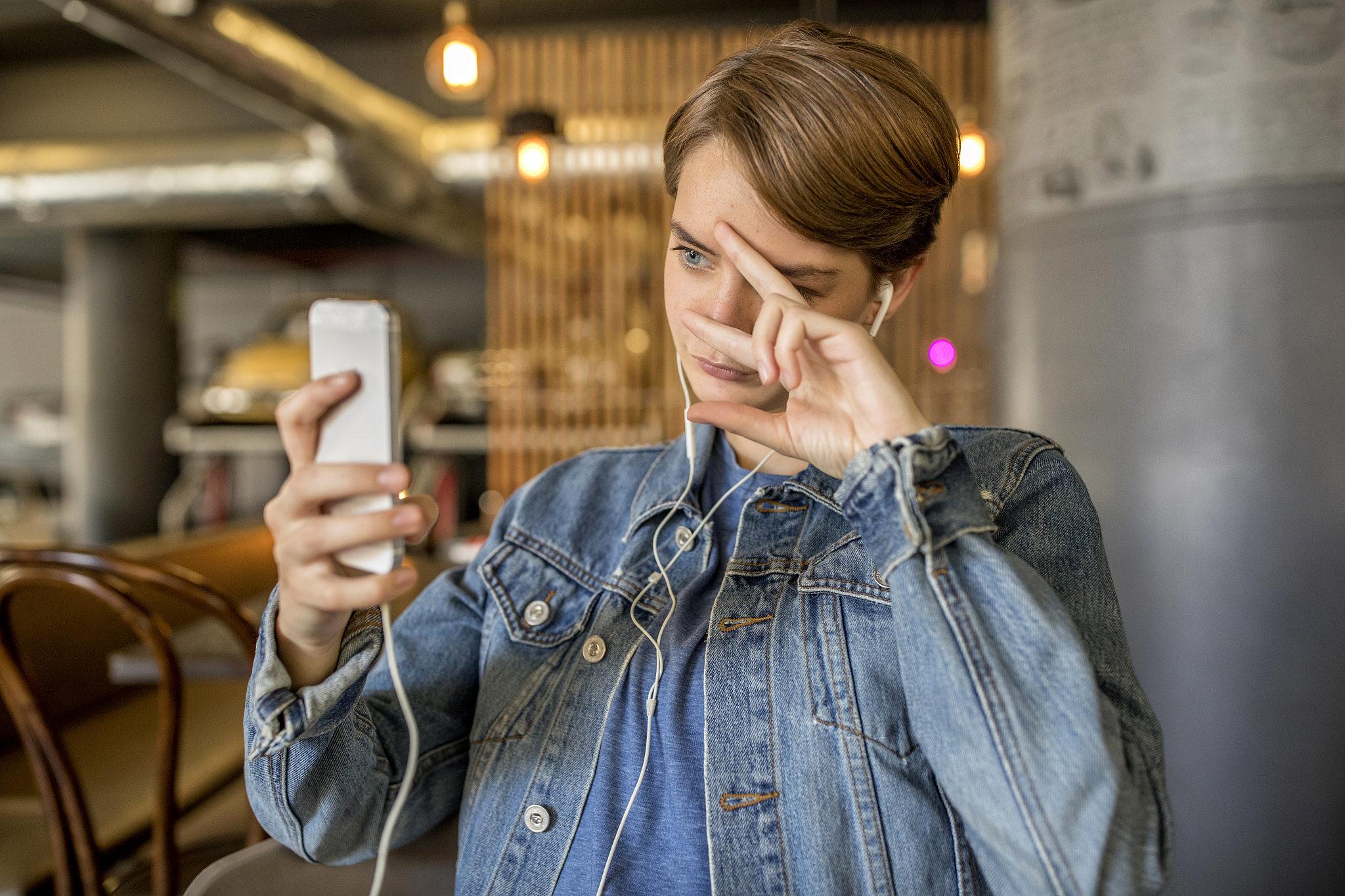 young woman with earbuds and smartphone in cafe gesturing