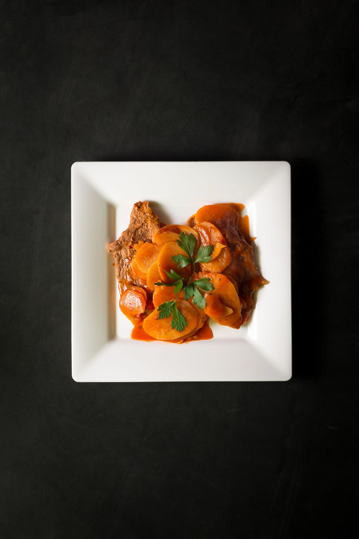 steak with carrots and potatoes on a square white plate on a bla