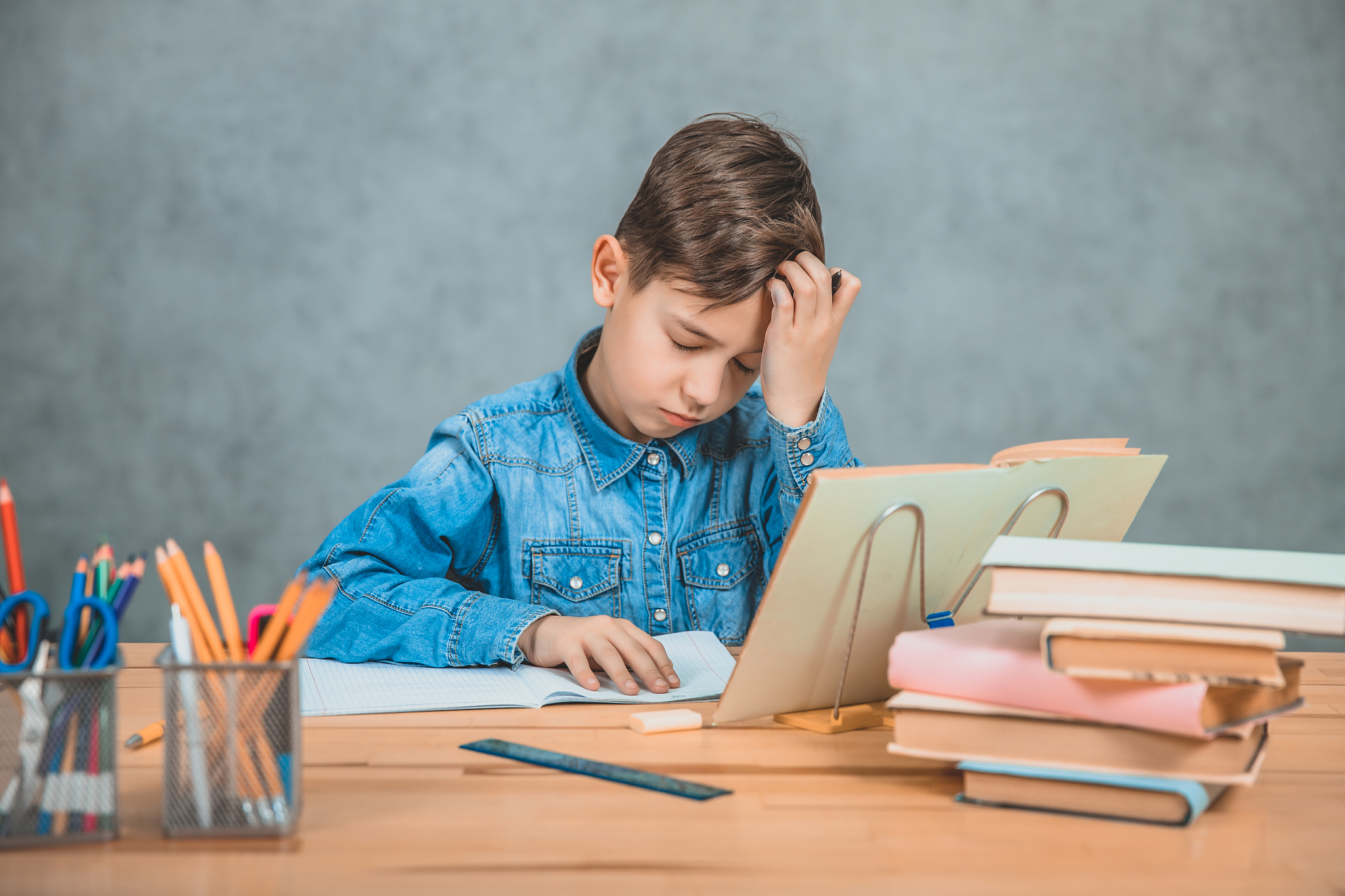 little tired schoolboy is sitting at the desk, overwhelmed with 