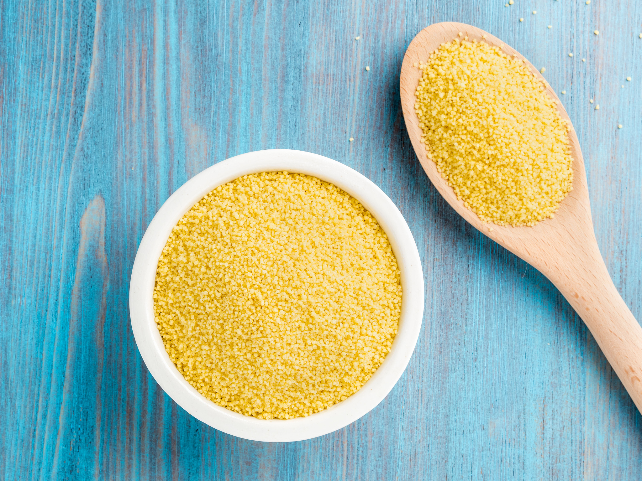 white bowl  and wooden spoon with raw couscous on blue wooden ba