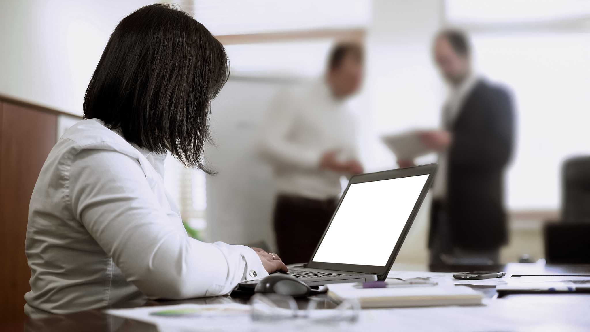 female employee typing minutes on computer in meeting, men discu