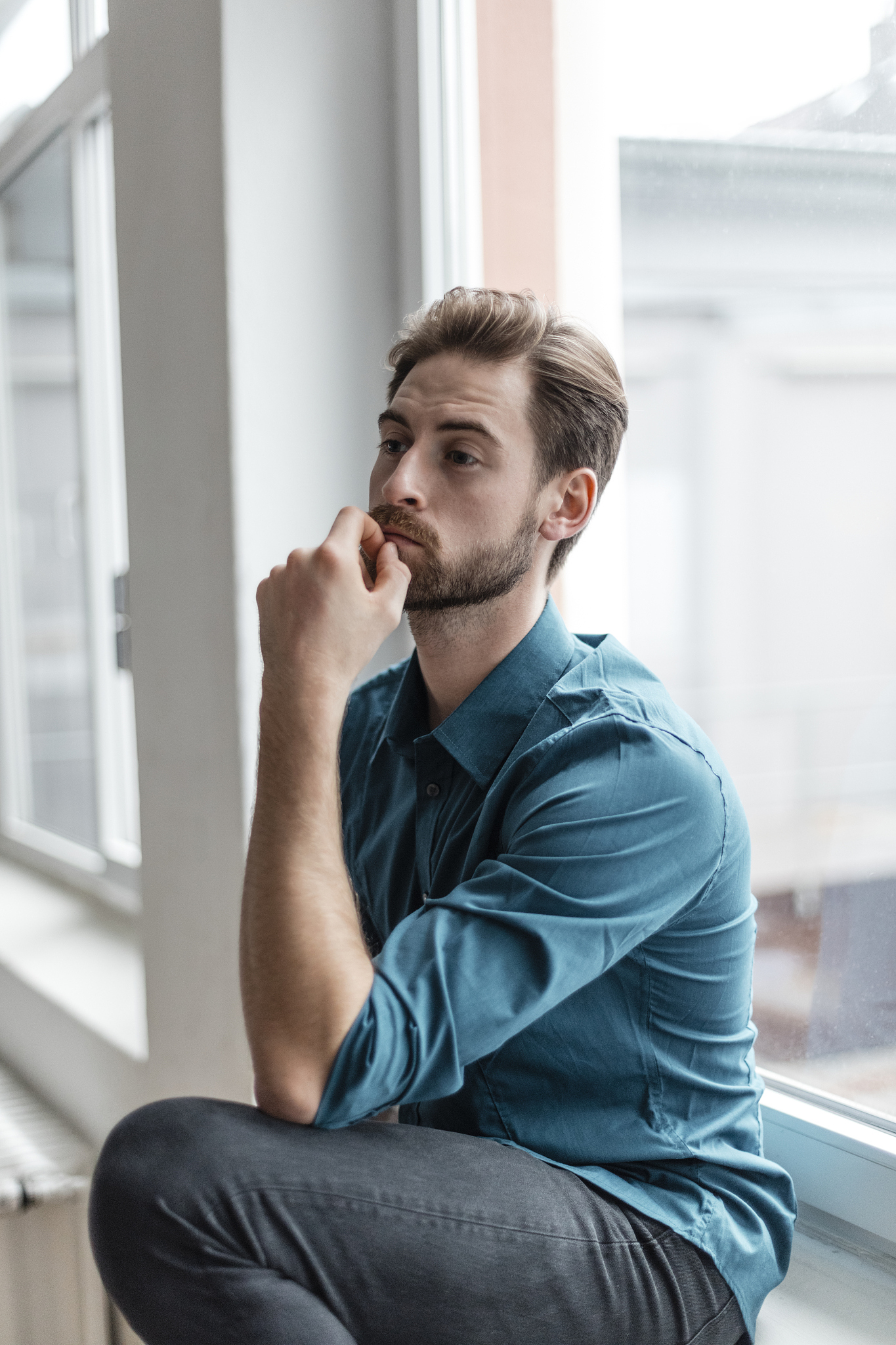 portrait of pensive young man sitting on window sill