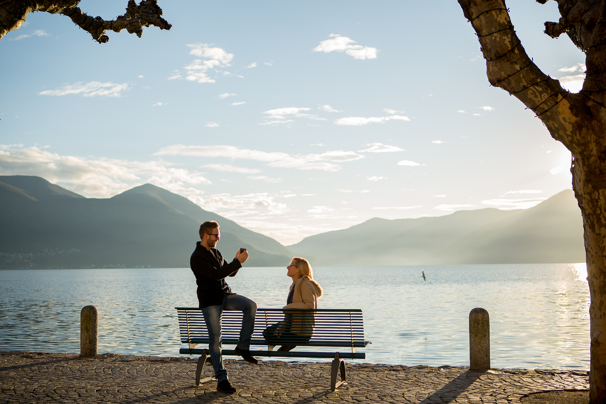 man takes picture of woman on bench, lake edge