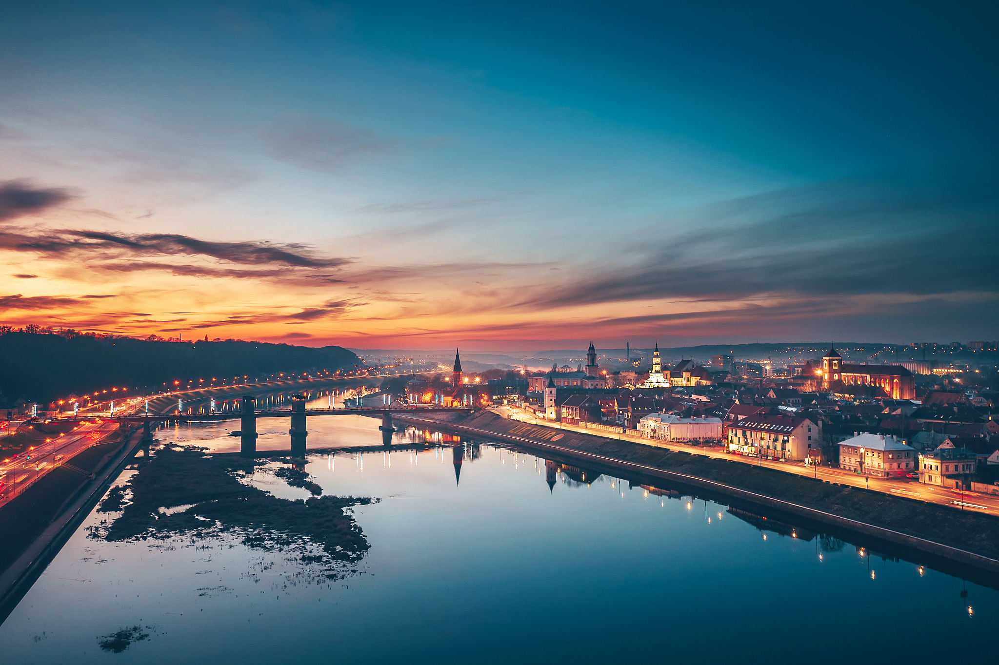 illuminated city by river against sky at sunset