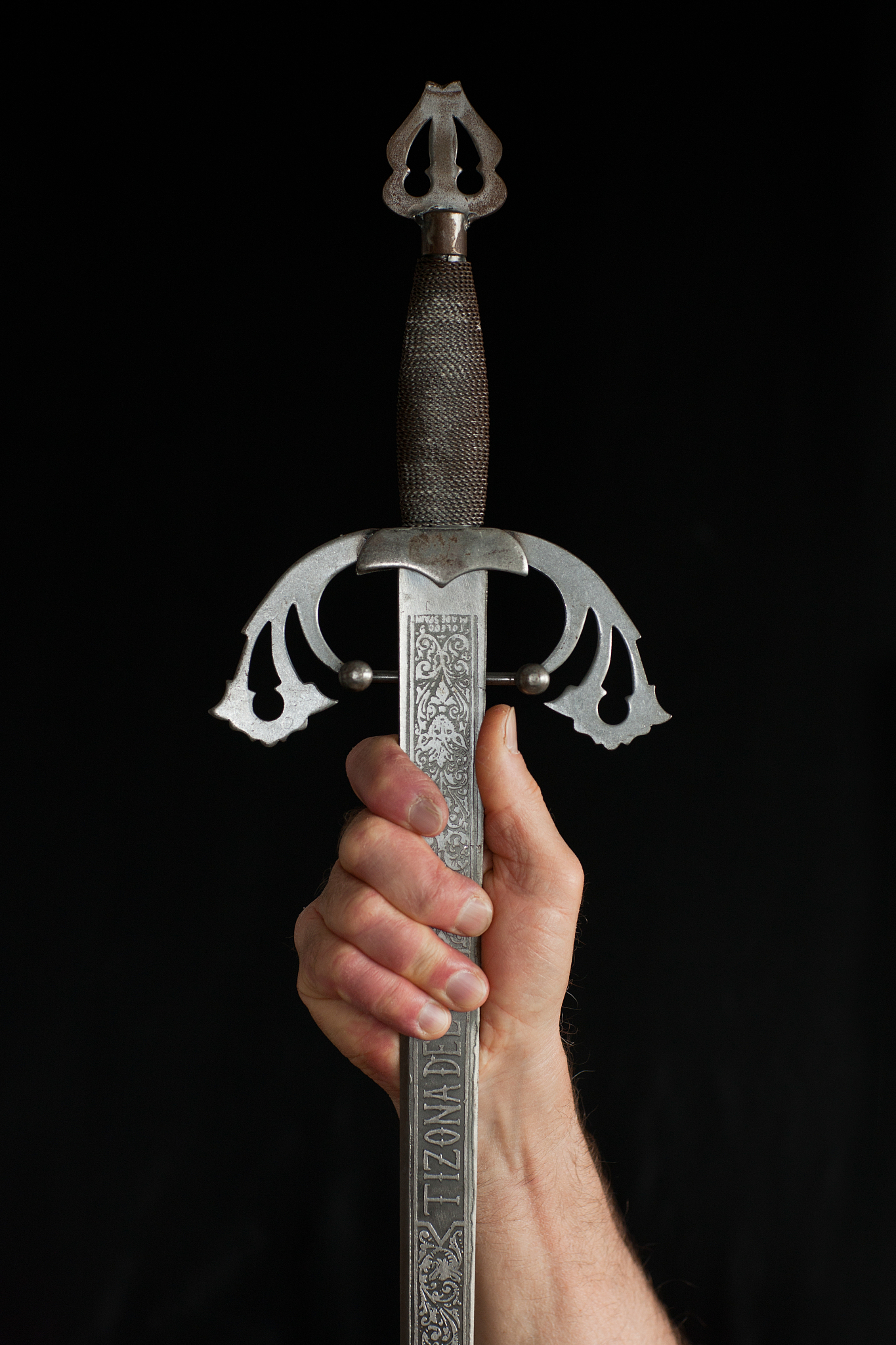 hand holding raised sword by blade, black background