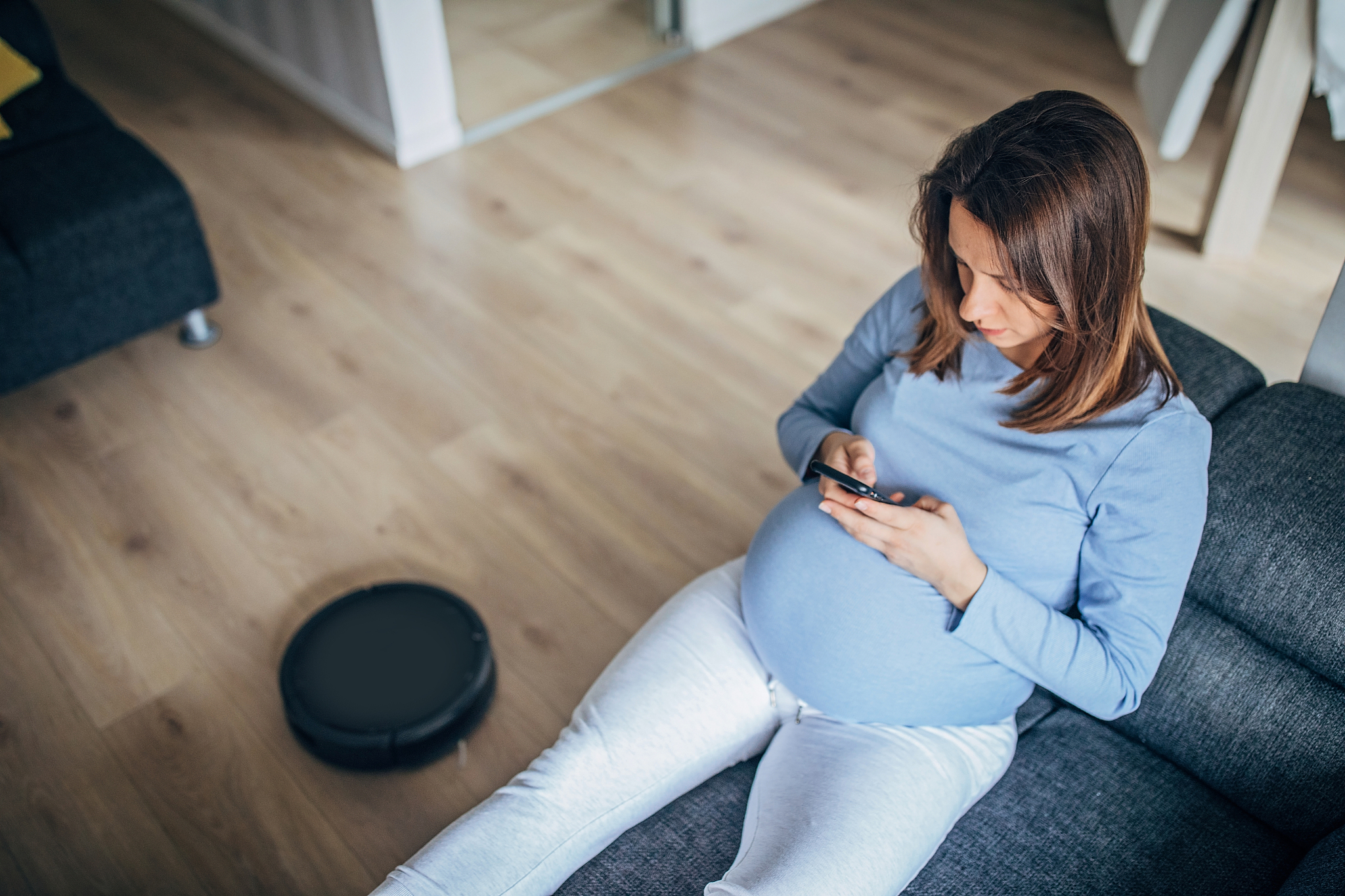 pregnant woman texting while robot vacuum cleaner cleaning floor