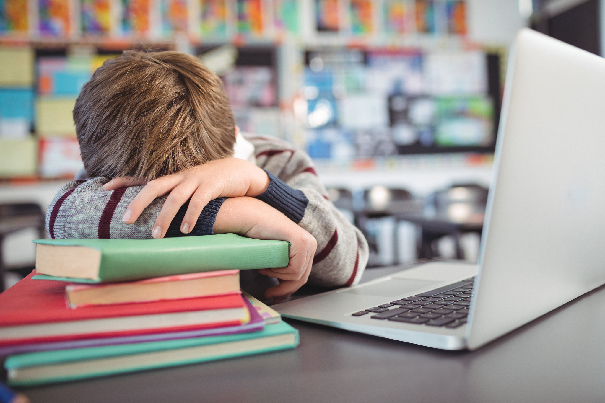 tired schoolboy resting on stack of book by laptop in classroom