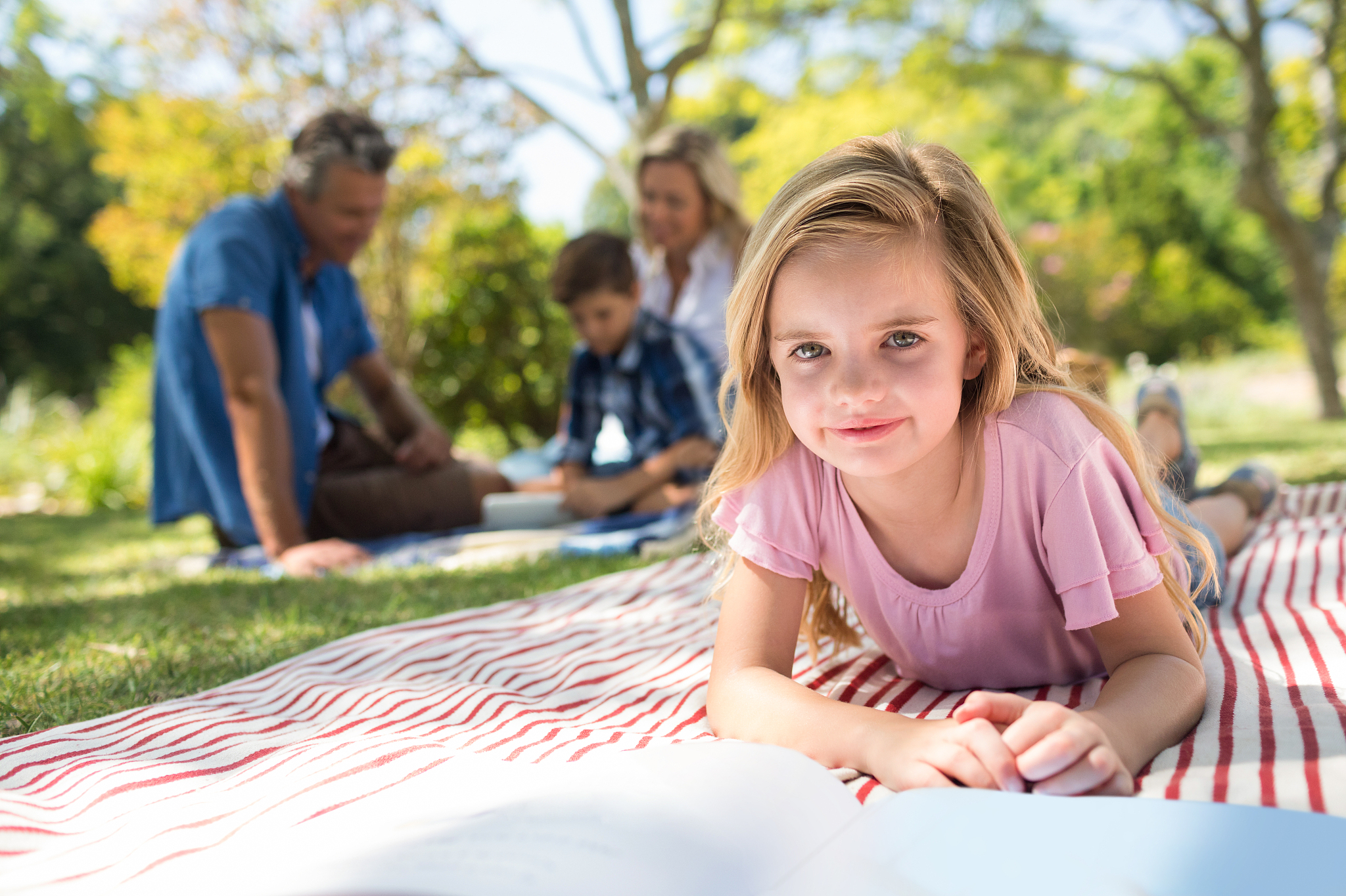 girl lying on blanket and reading book while family sitting in b
