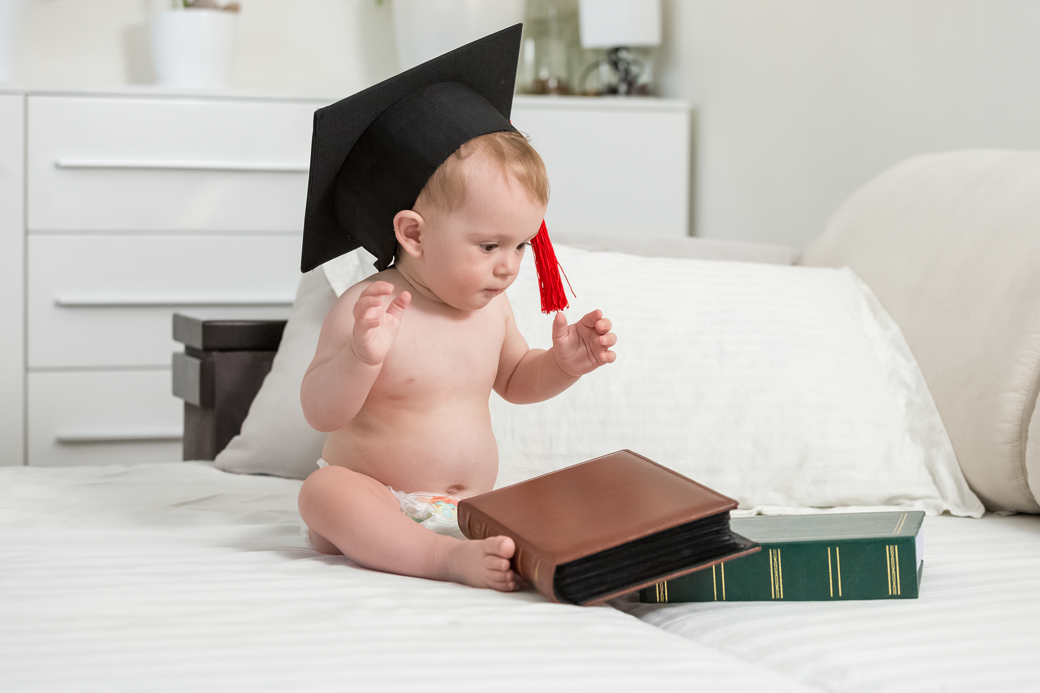 baby wearing black graduation cap looking at stack of books