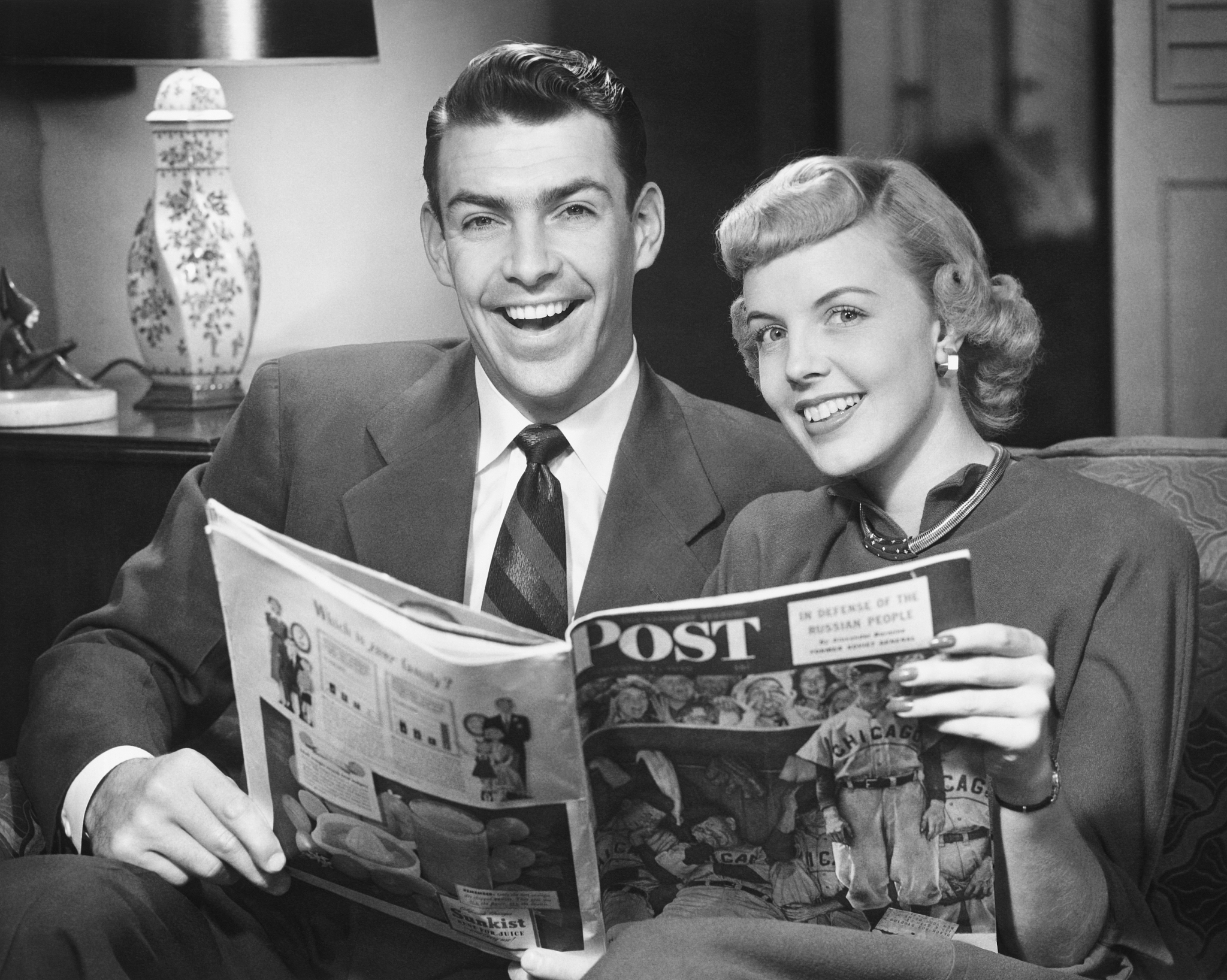 couple sitting on sofa, holding magazine, (b&w), portrait
