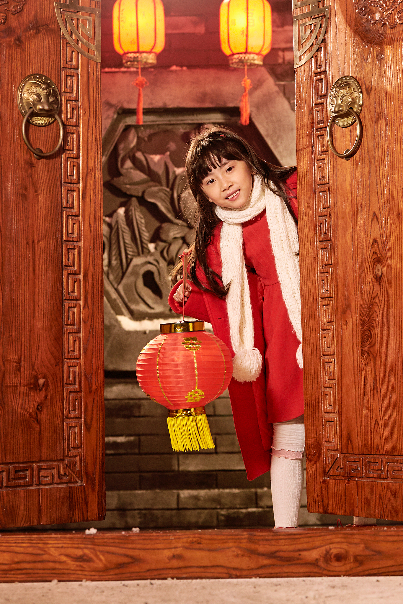 图中,一位红衣小女孩手捧红灯笼,笑容满面,身后是家家户户贴春联,挂
